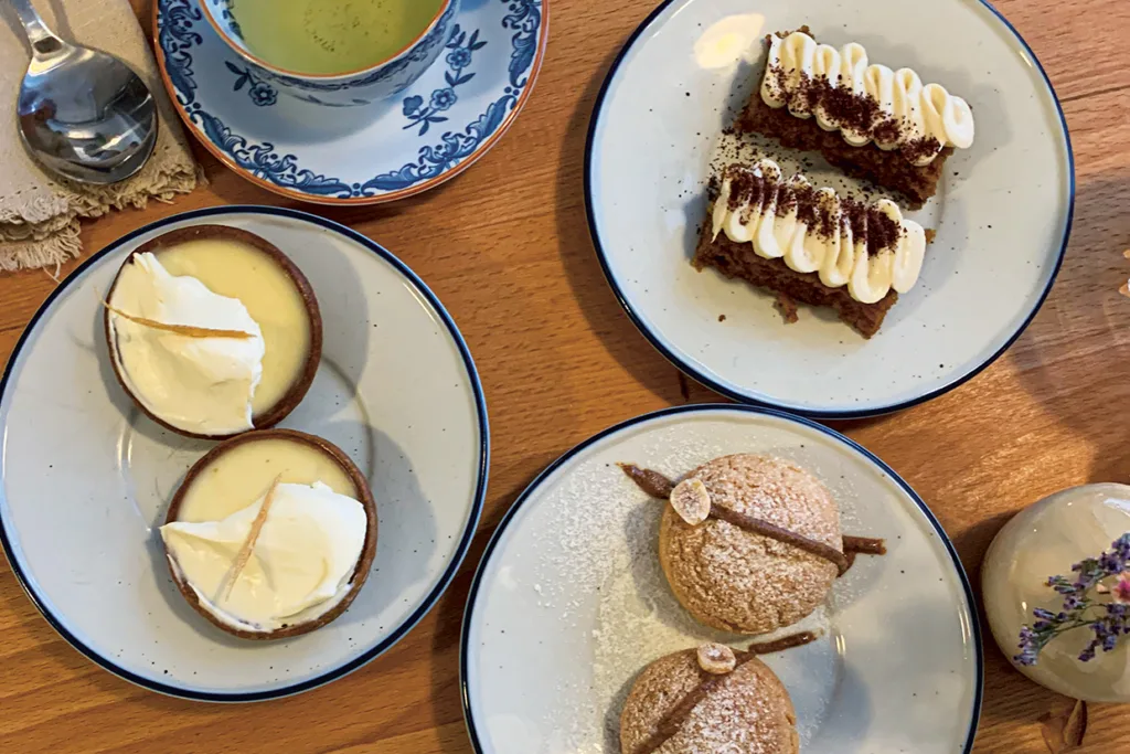 Ett bord dukat med desserter: två citronpajer toppade med grädde, två gräddfyllda bakelser pudrade med florsocker, två morotskakor med glasyr och en kopp grönt te på en träskiva.