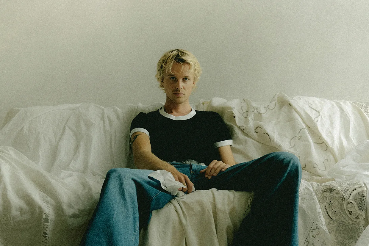 Isak Danielson med kort blont hår sitter i en vit soffa med vita lakan, klädd i en svartvit t-shirt och blå jeans, och tittar rakt in i kameran med ett neutralt uttryck.