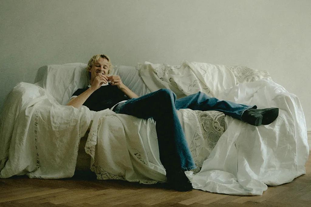 Isak Danielson med kort blont hår slappnar av i en soffa täckt av vita lakan, ler och håller i en drink, med ett ben utsträckt och det andra böjt, i ett minimalt inrett rum med trägolv.