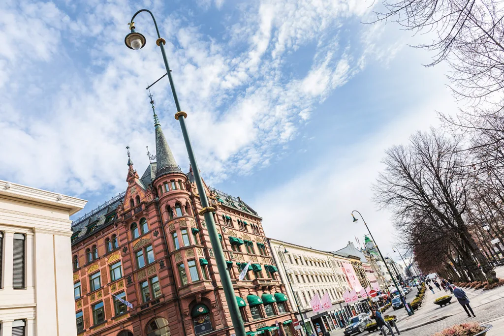 En historisk röd tegelbyggnad med utsmyckade spiror och gröna markiser står på en livlig stadsgata under en blå himmel med spridda moln. Människor går längs trottoaren och bilar är parkerade vid trottoarkanten.
