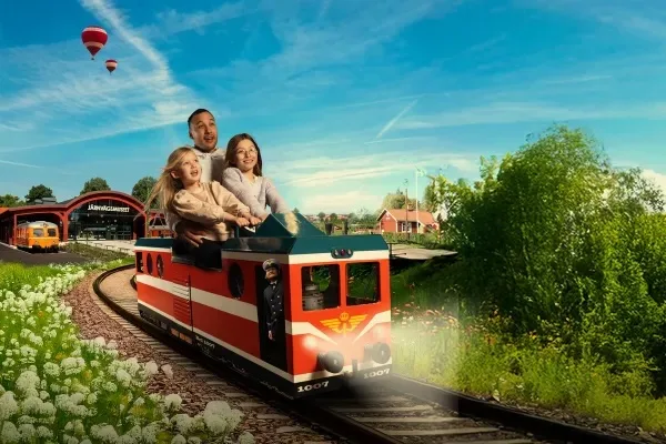 En leende familj på fyra personer åker ett litet, rött och vitt friluftståg en solig dag, omgiven av grönska och blommor, med luftballonger och en station i bakgrunden.