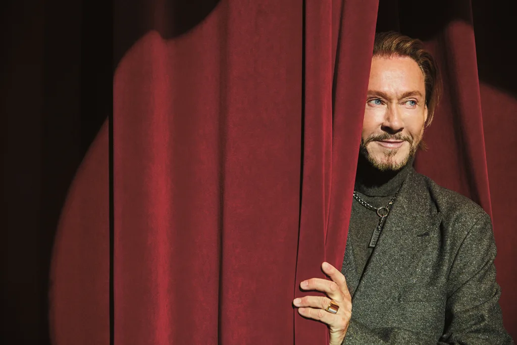 Christer Lindarv med vältrimmat skägg och mustasch, klädd i mörk rock, tittar fram bakom en röd teaterridå, ler och håller ridån med ena handen. Dramatisk ljussättning framhäver hans ansikte.
