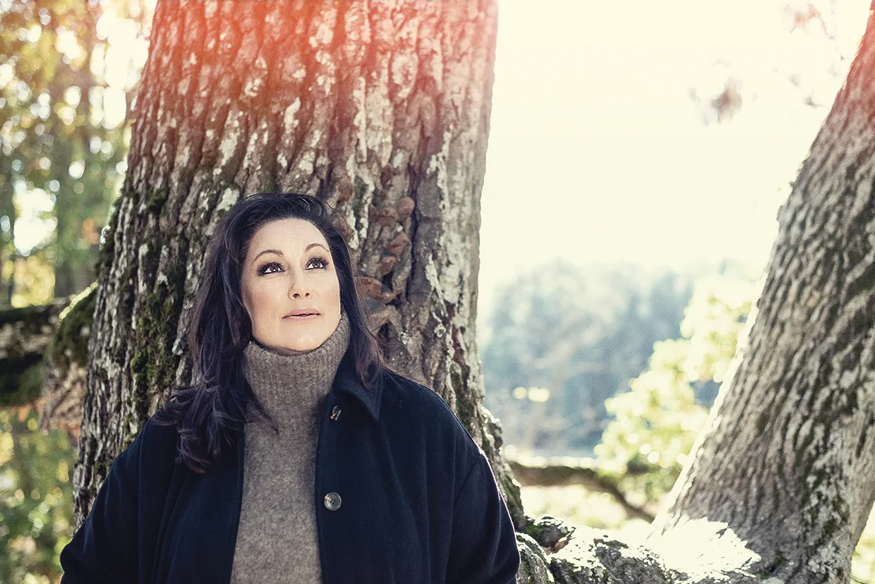 Lisa Nilsson med långt mörkt hår, klädd i mörk kappa och höghalsad tröja, står utomhus lutad mot ett stort träd och tittar tankfullt upp. Solljuset silas genom grenarna i bakgrunden.