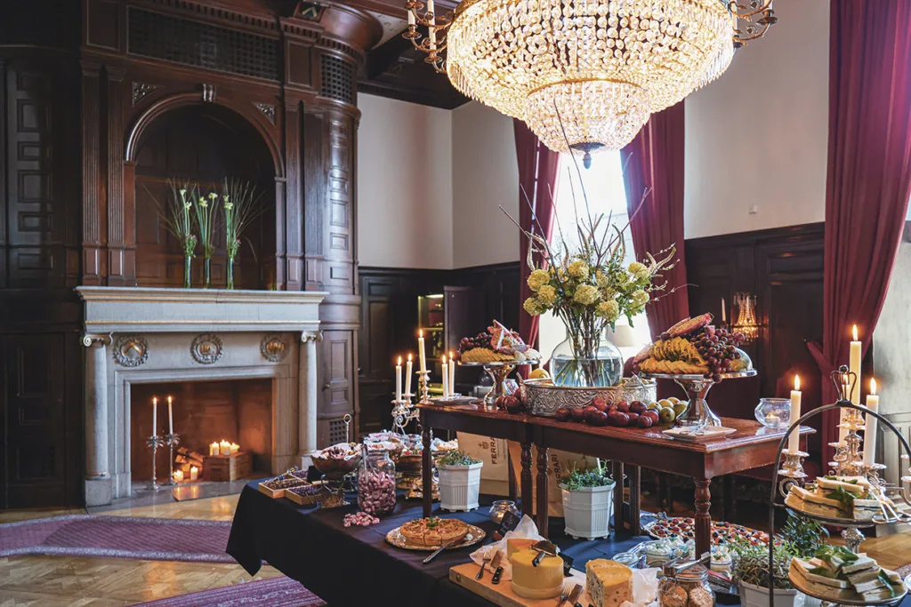 En påkostad matsal med ljuskrona, öppen spis och höga fönster. Ett långbord är fyllt med eleganta rätter, frukt, bakverk och blomsterdekorationer, vilket skapar en inbjudande och lyxig atmosfär.