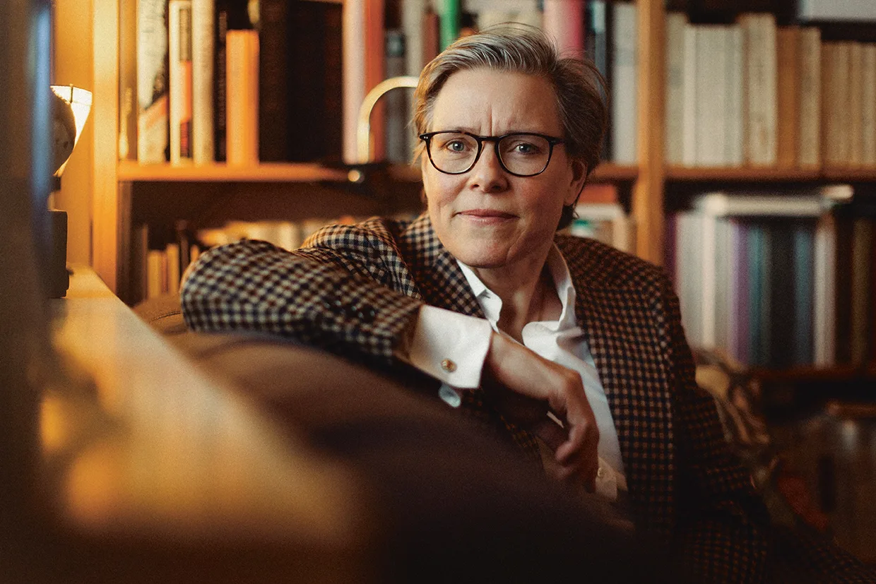 Lena Andersson med kort grått hår och glasögon, klädd i rutig kavaj och vit skjorta, sitter i en soffa i ett mysigt rum fyllt med bokhyllor och tittar eftertänksamt in i kameran.