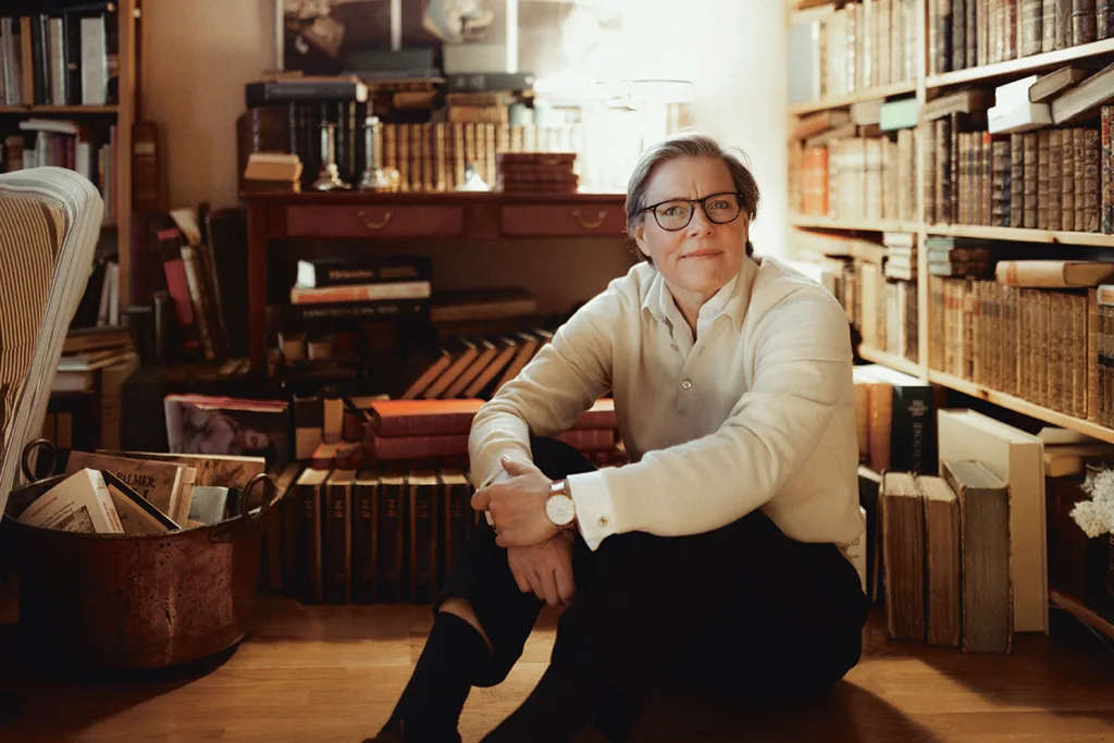 Lena Andersson med kort hår och glasögon sitter på golvet, leende och omgiven av bokhögar och hyllor fyllda med böcker i ett mysigt, varmt upplyst rum.