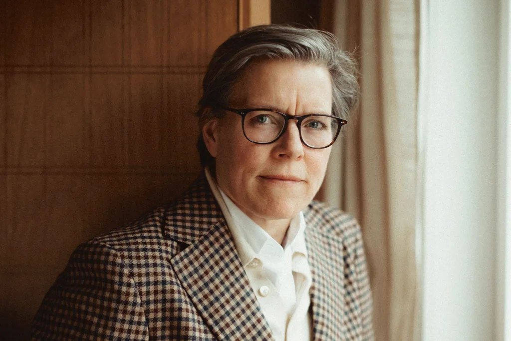 Lena Andersson med kort grått hår och glasögon, klädd i en rutig kavaj över en vit skjorta, står inomhus vid ett fönster med beige gardiner och tittar direkt in i kameran med ett neutralt ansiktsuttryck.