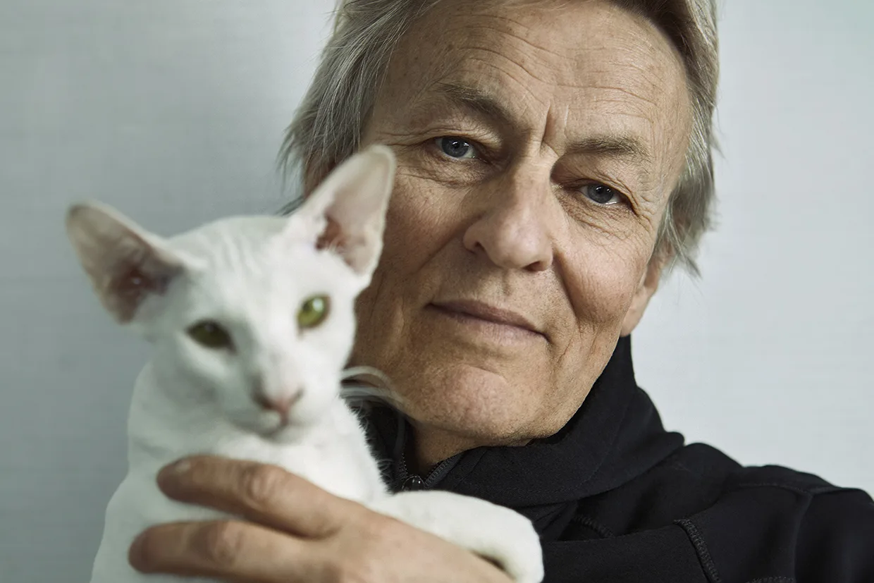 Lars Lerin håller en vit, smal katt med stora öron nära ansiktet, båda tittar mot kameran mot en slät bakgrund.