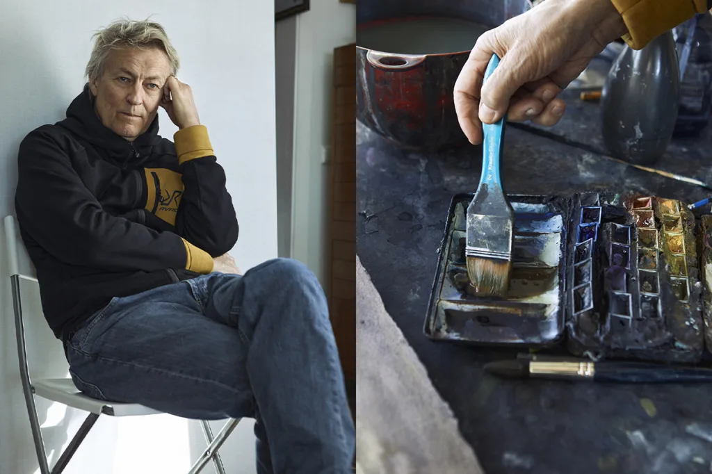 Lars Lerin med ljust hår sitter på en vit stol och vilar huvudet på ena handen. Bredvid syns i närbild en hand som doppar en pensel i en akvarellpalett på ett rörigt konstbord.