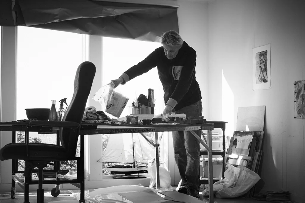Lars Lerin står vid ett rörigt bord i ett ljust rum och håller på med ett konstprojekt. Konstmaterial och papper täcker bordet och olika konstverk och föremål är synliga runt om i rummet. Scenen är i svartvitt.