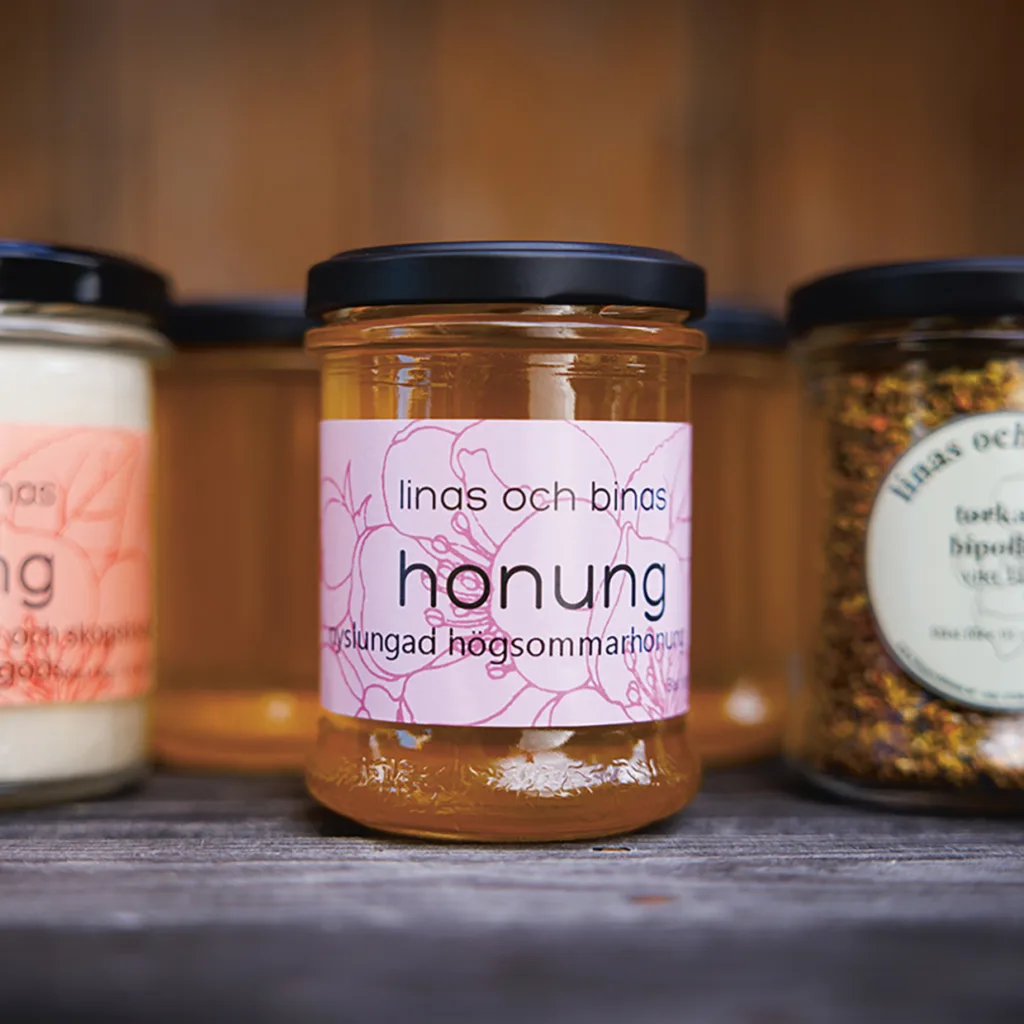 En glasburk med honung med en rosa blommig etikett med texten linas och binas honung står på en träyta, omgiven av andra burkar. Honungen är gyllene och bakgrunden är mjukt suddig.