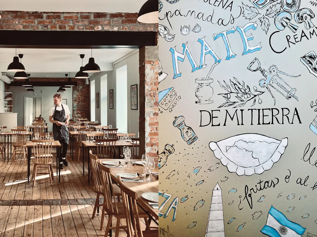 En servitör står i en ljus, rustik restaurang med träbord och synliga tegelväggar. Bredvid finns en väggmålning med handritade illustrationer och spanska ord om det argentinska köket, som mate, empanadas och de mi tierra.