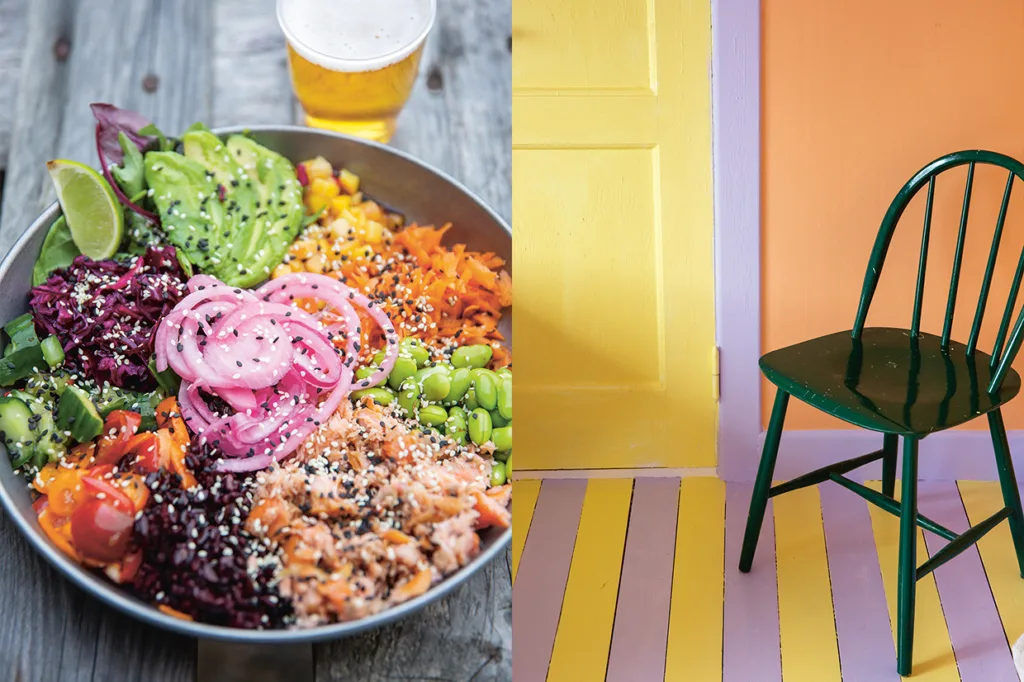 En stor skål med sallad med avokado, picklad lök, tomater och sesamfrön står bredvid ett glas öl på ett träbord; bredvid står en grön stol på ett gul- och lila randigt golv mot en gul- och orange vägg.