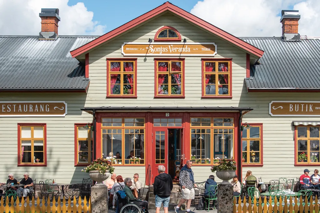 Människor sitter utanför en charmig, ljusgrön träbyggnad med rödmålade fönster, märkt "Sonjas Veranda". Skyltar för en restaurang och en butik syns. Scenen är livlig och solig med en blå himmel.