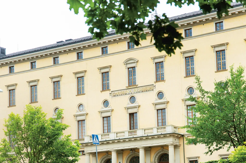 En beige, historisk byggnad med höga välvda fönster och dekorativa pelare, delvis inramad av gröna träd. Skylten CAROLINA REDIVIVA syns ovanför entrén.