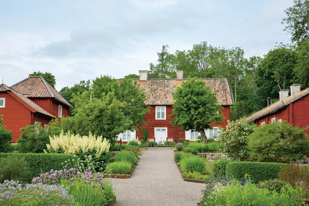 En grusgång leder genom en lummig trädgård med blommande blommor och välskötta buskar till ett rött trähus med vita lister och tegeltak, omgivet av träd och grönska.