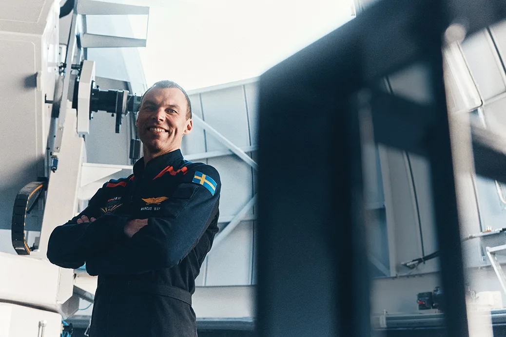 Marcus Wandt i mörk uniform med en svensk flagglapp står inomhus med armarna i kors och ler framför stor teknisk utrustning och moderna industribyggnader.