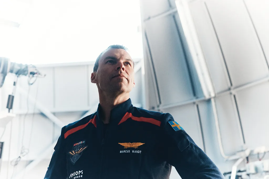 Marcus Wandt i mörk jumpsuit med flygplansmärken och en svensk flagga på ärmen står inomhus och tittar uppåt med självsäker blick. Starkt ljus skiner från vänster.