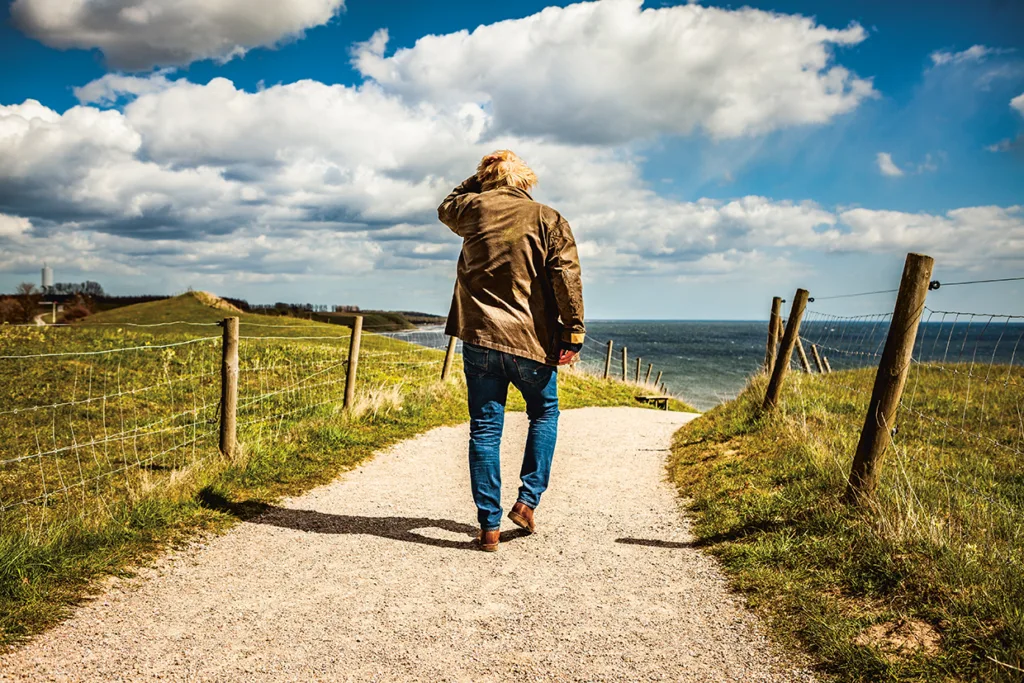 Rolf Lassgård i brun jacka och jeans går ensam på en grusväg längs en gräsbevuxen kustlinje under en delvis molnig himmel, med havet synligt i fjärran.