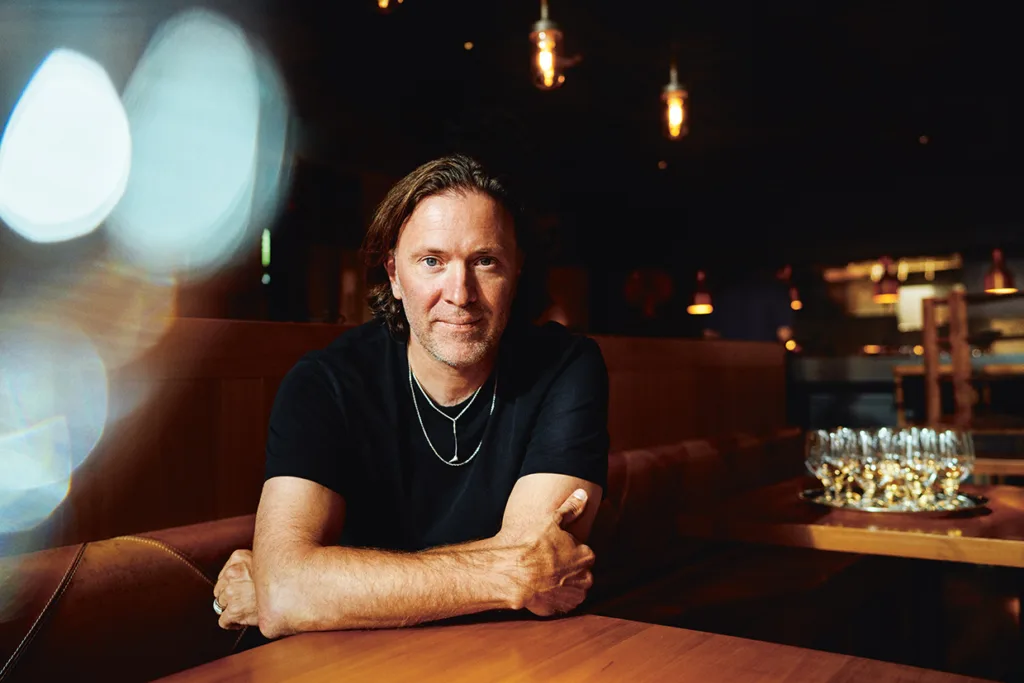 Niklas Ekstedt med axellångt brunt hår, svart t-shirt och silverhalsband sitter vid ett träbord i en svagt upplyst restaurang och ler med armarna i kors på bordet. Glasögon syns i bakgrunden.