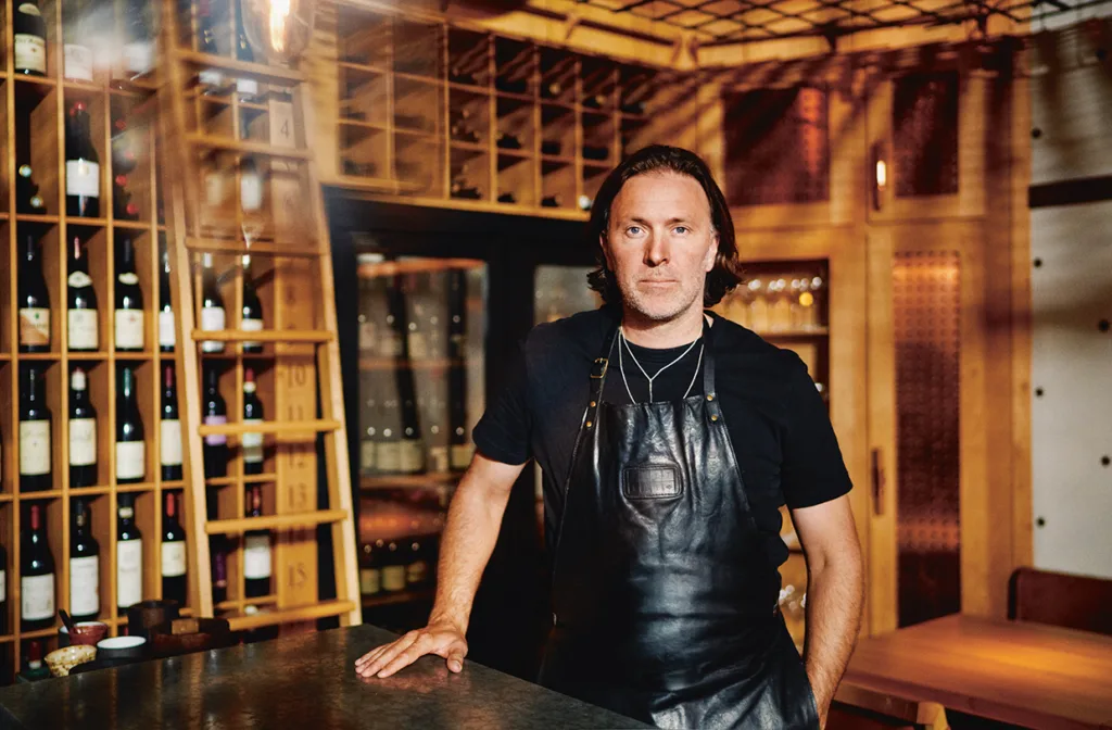 Niklas Ekstedt i svart förkläde står bakom en disk i en vinkällare eller restaurang, med hyllor av vinflaskor i bakgrunden och varm belysning som fyller utrymmet.