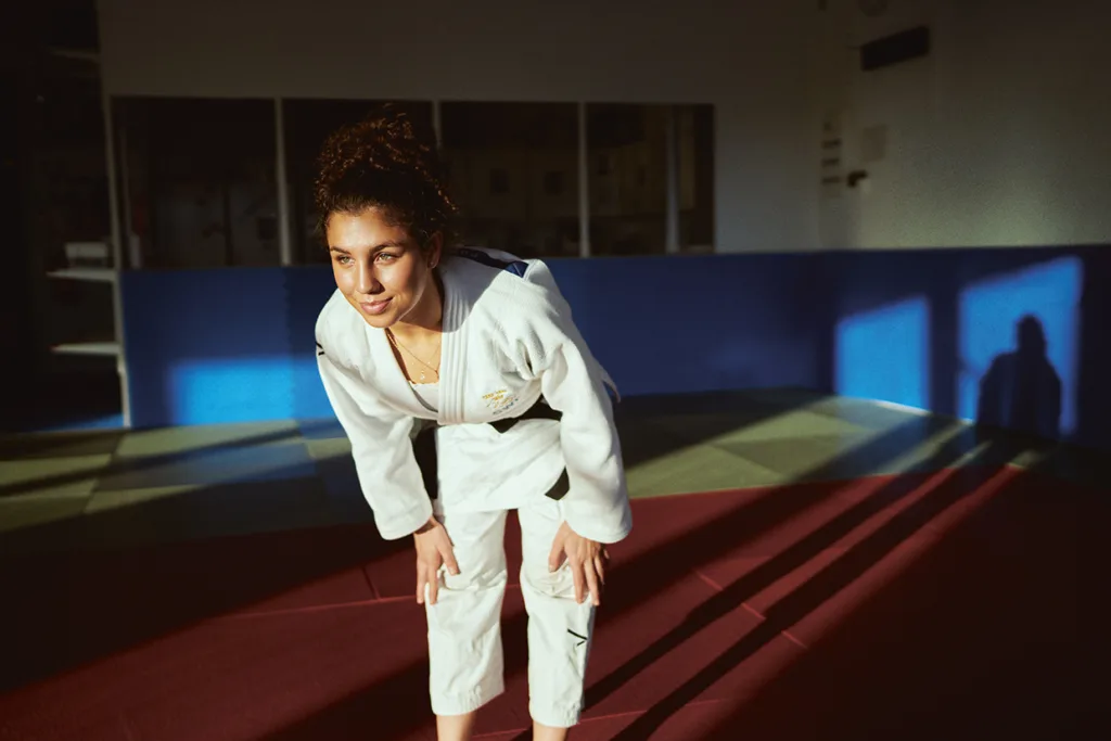 Tara Babulfath i vit judodräkt står på en röd matta i ett solbelyst rum, lutar sig framåt med händerna på knäna och ler. Blått och gult ljus kastar skuggor på vägg och golv.