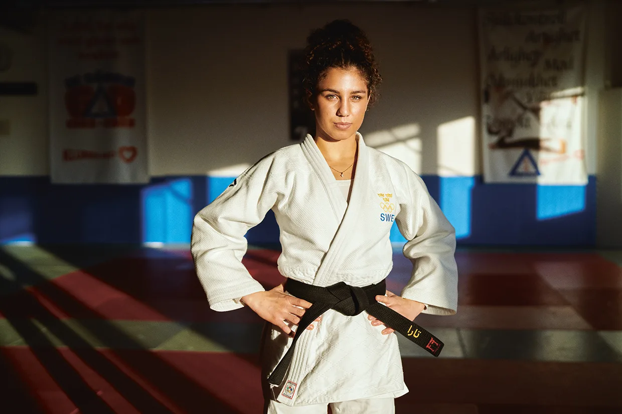 Tara Babulfath i vit judogi med svart bälte står självsäkert med händerna på höfterna i en dojo, solljuset flödar in från sidan. Banderoller hänger på väggen i bakgrunden.