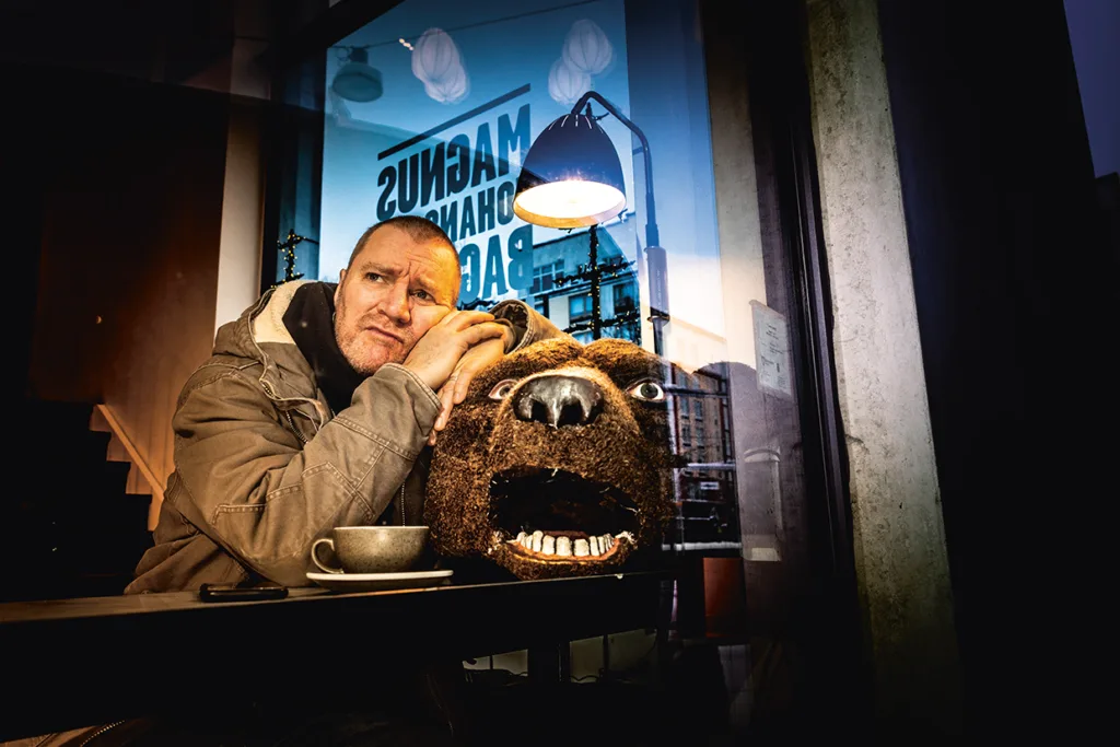 Magnus Krepper i luvjacka sitter vid ett cafébord med en kopp och vilar huvudet på ett stort huvud i björndräkt. I fönstret bakom honom syns en lampa och stadsreflexer.