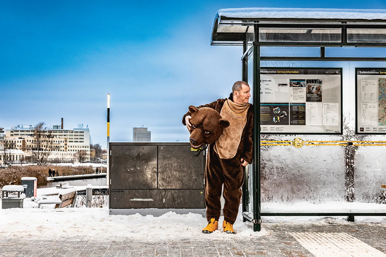 Magnus Krepper i björndräkt, med björnhuvudbonad, står vid en snöig busshållplats bredvid en informationstavla en klar vinterdag, med byggnader i bakgrunden.