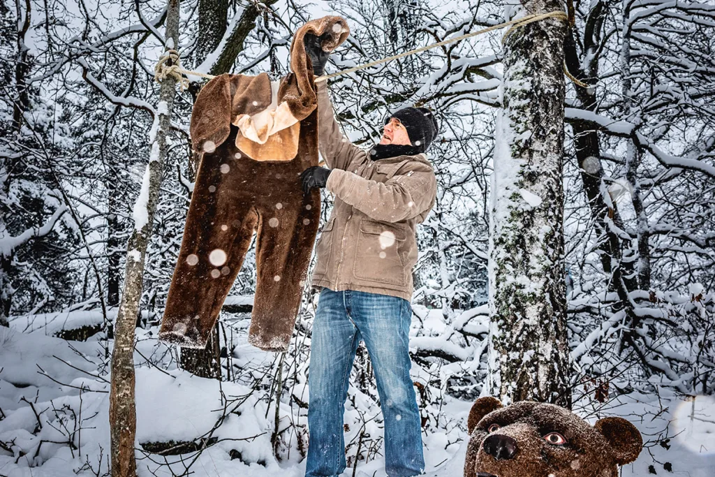 Magnus Krepper i vinterkläder hänger upp en stor brunbjörnsdräkt på en lina mellan snöiga träd i en skog. Snön täcker marken och grenarna, och björndräktens huvud vilar i närheten.