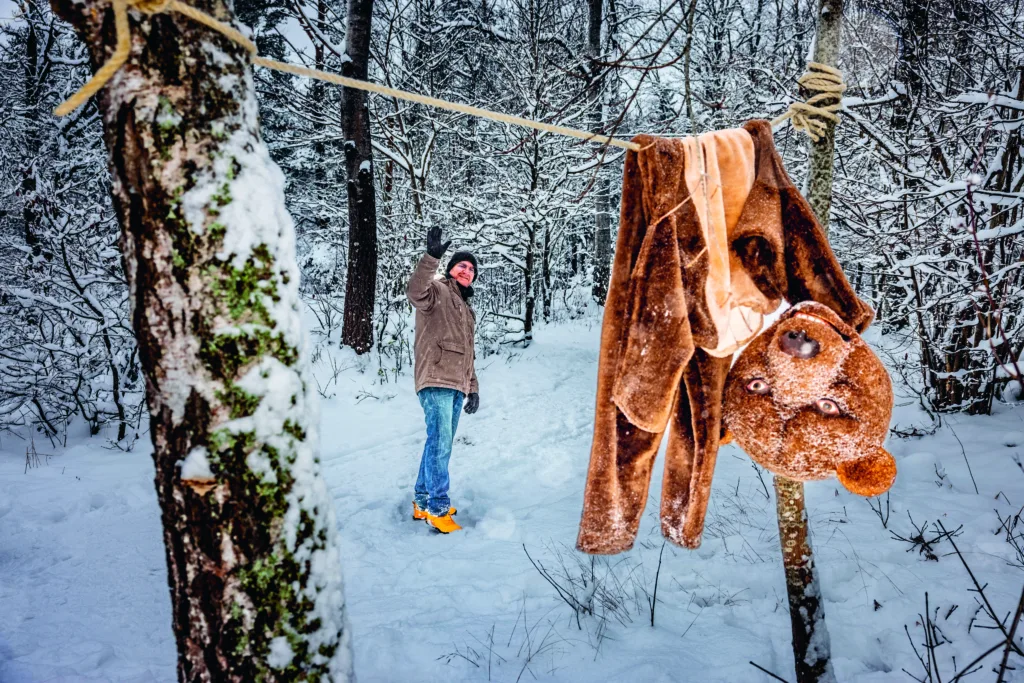 Magnus Krepperi vinterkläder vinkar när han står i en snöig skog; i förgrunden hänger en björndräkt upp och ner på ett rep som är bundet mellan två träd.