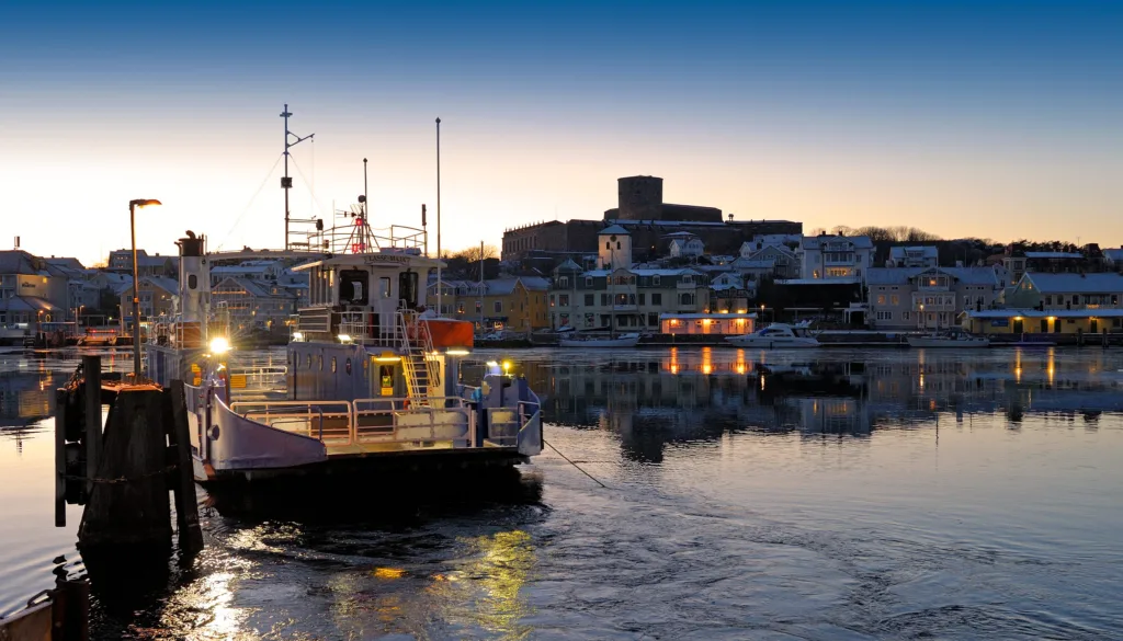 En färja med ljus på ligger i en lugn hamn i skymningen, med färgglada byggnader och båtar längs vattnet och en stor fästning på en kulle i bakgrunden.