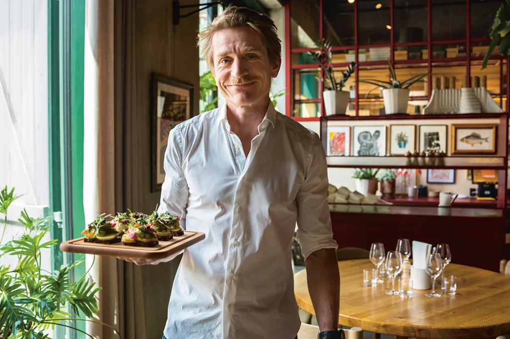 En leende man i vit skjorta står på en restaurang och håller i ett trätallrik med aptitretare; bakom honom finns ett dukat bord, krukväxter och inramade konstverk på röda hyllor.