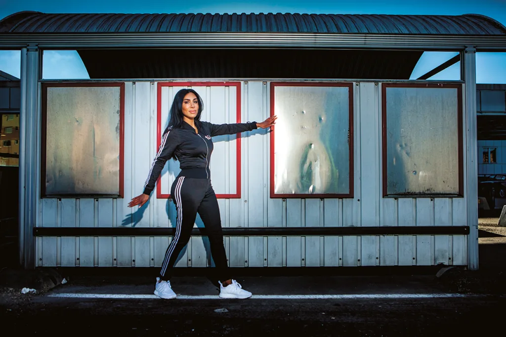 En kvinna i svart träningsoverall och vita sneakers poserar med utsträckta armar framför en metallvägg med fyra frostade fönster, utomhus en solig dag.