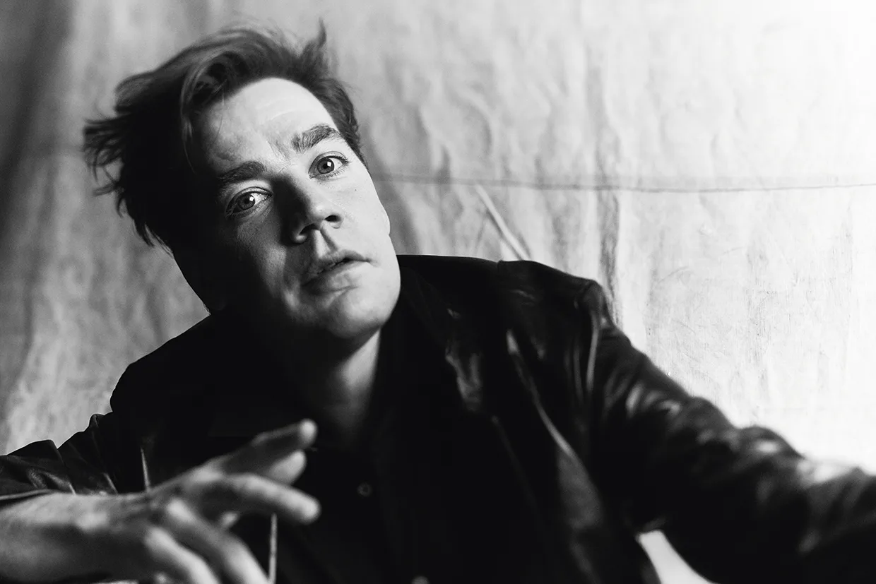 Svartvitt foto av Pelle Almqvist med rufsigt hår, klädd i mörk jacka, som tittar intensivt in i kameran med ena handen utsträckt mot en strukturerad, ljus bakgrund.