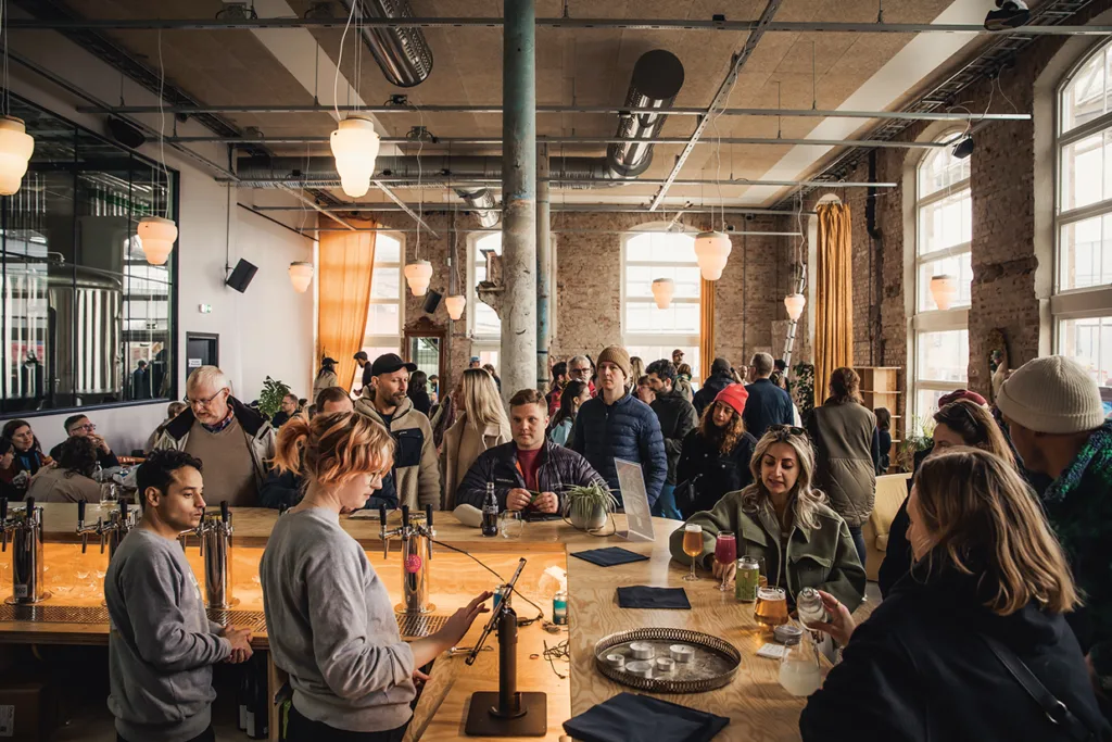 Ett livligt café med stora fönster och industriell inredning, fyllt av människor som pratar, dricker och umgås; personalen förbereder drinkar bakom en trädisk under hängande lampor.