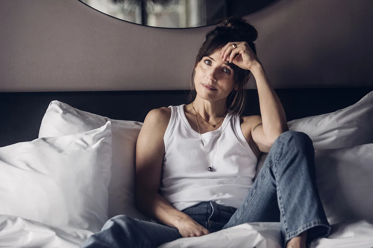Tuva Novotny i vitt linne och jeans sitter på en säng med vita kuddar, vilar armbågen på knät och handen på pannan och ser eftertänksam och avslappnad ut.