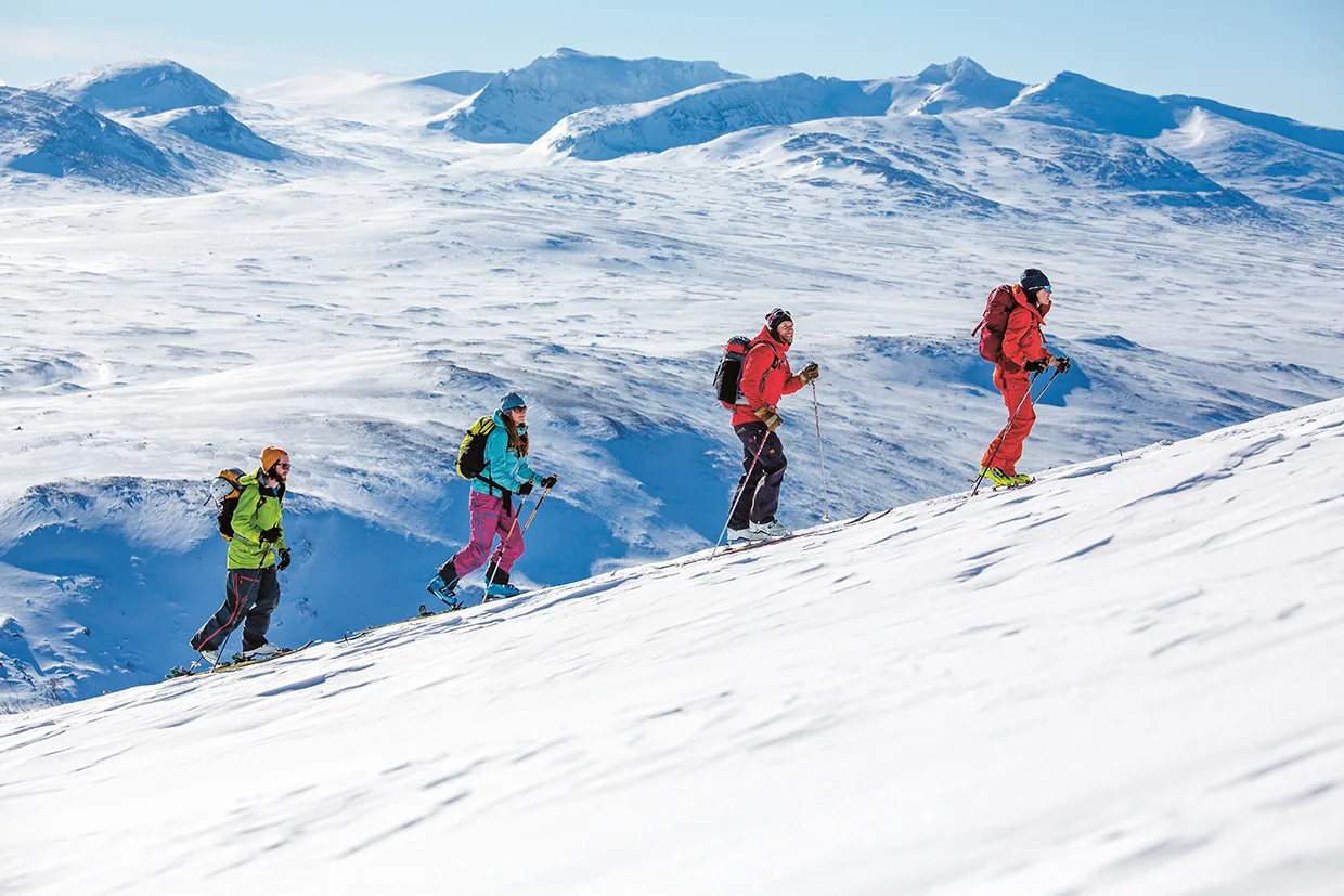 Fyra personer med färgglada vinterkläder och ryggsäckar åker skidor uppför en snöig bergssluttning, med snötäckta bergstoppar och en klarblå himmel i bakgrunden.