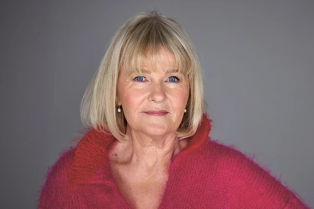 Sissela Kyle med rakt blont hår och lugg bär en ljusrosa lurvig tröja, står mot en vanlig grå bakgrund och tittar självsäkert in i kameran.