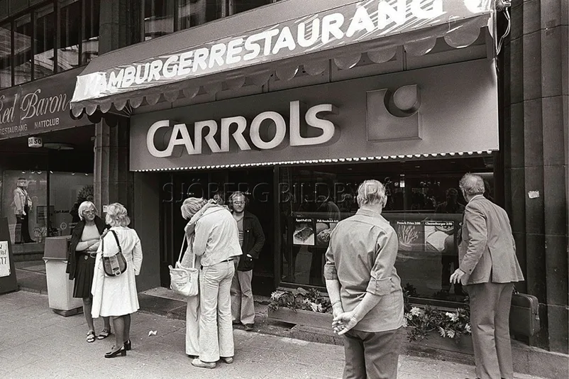 En grupp människor står och samtalar utanför en retro Carrols hamburgerrestaurang, med en stor skylt ovanför entrén, på en gata i en stad. Bilden är i svartvitt.