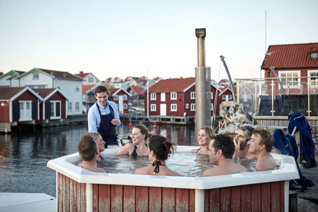 En grupp människor kopplar av i en utomhusbadtunna i trä vid vattnet, skrattar och pratar, medan en man i förkläde står bredvid dem. I bakgrunden syns röda och vita hus.