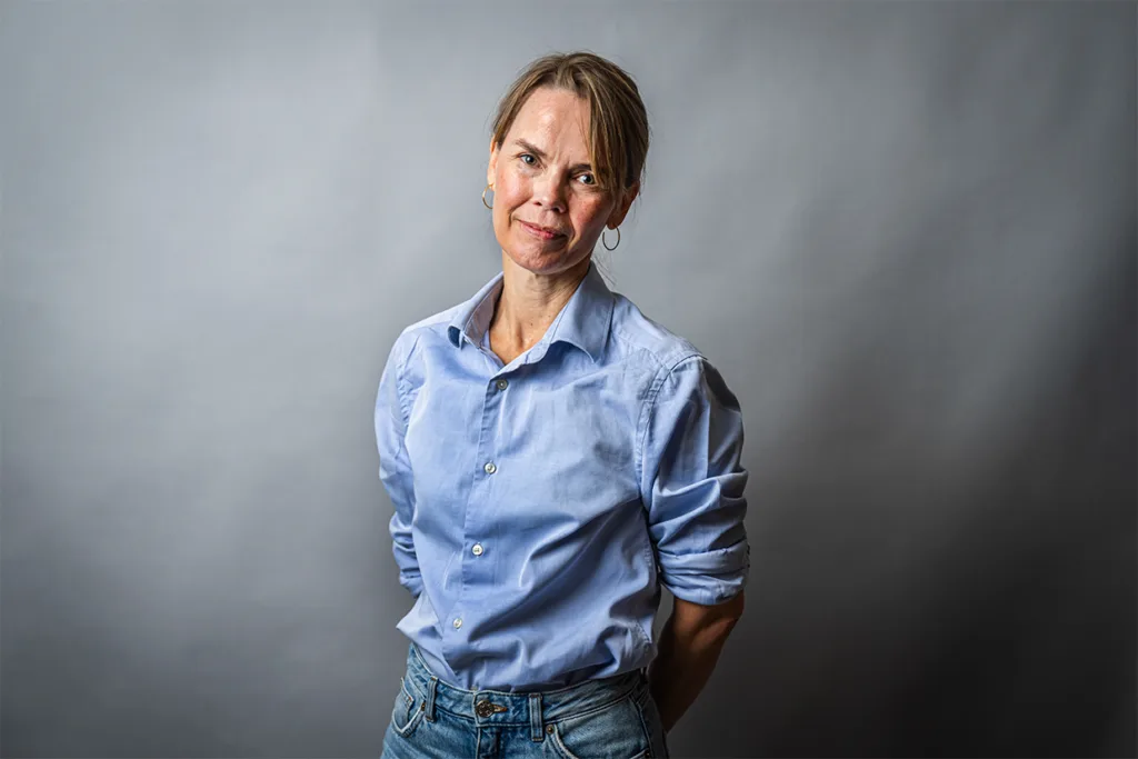 Stina Hindström med ljusbrunt hår, klädd i ljusblå skjorta med knappar och jeans, står mot en grå bakgrund och tittar in i kameran med ett litet leende och händerna bakom ryggen.
