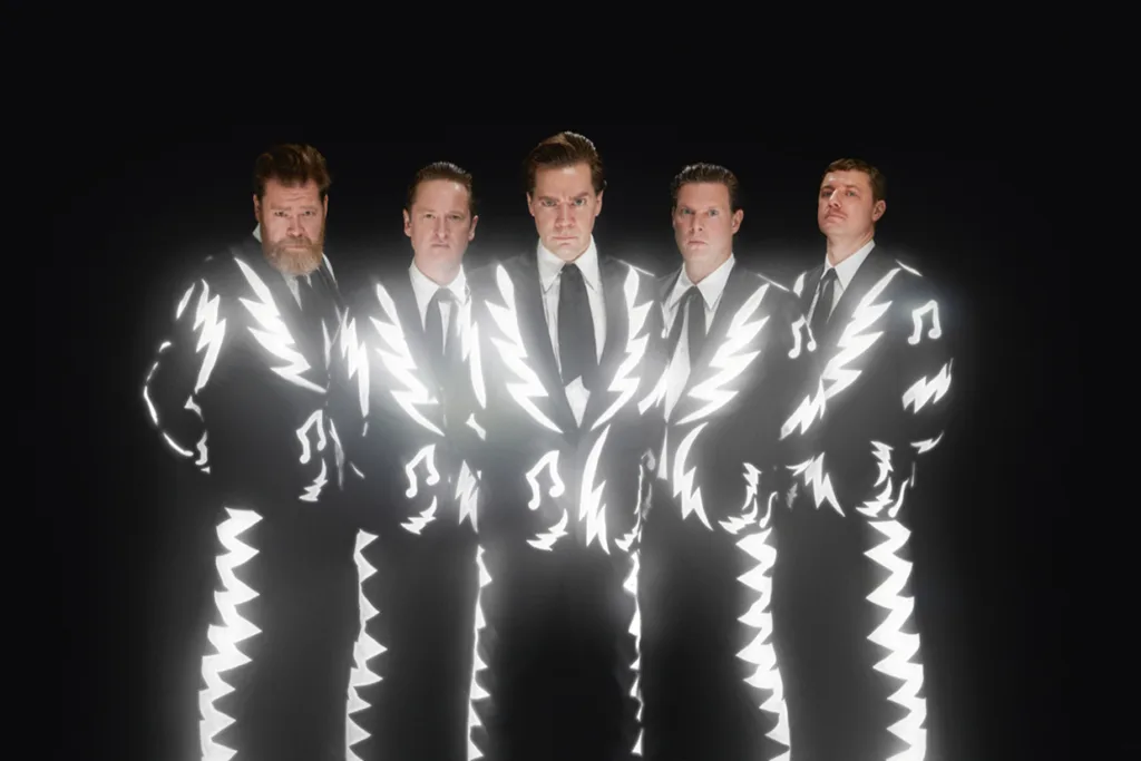 The Hives står sida vid sida mot en svart bakgrund. Deras kostymer har ljusa, lysande vita sicksack- och musiknotsmönster, vilket skapar en elektrisk, upplyst effekt.