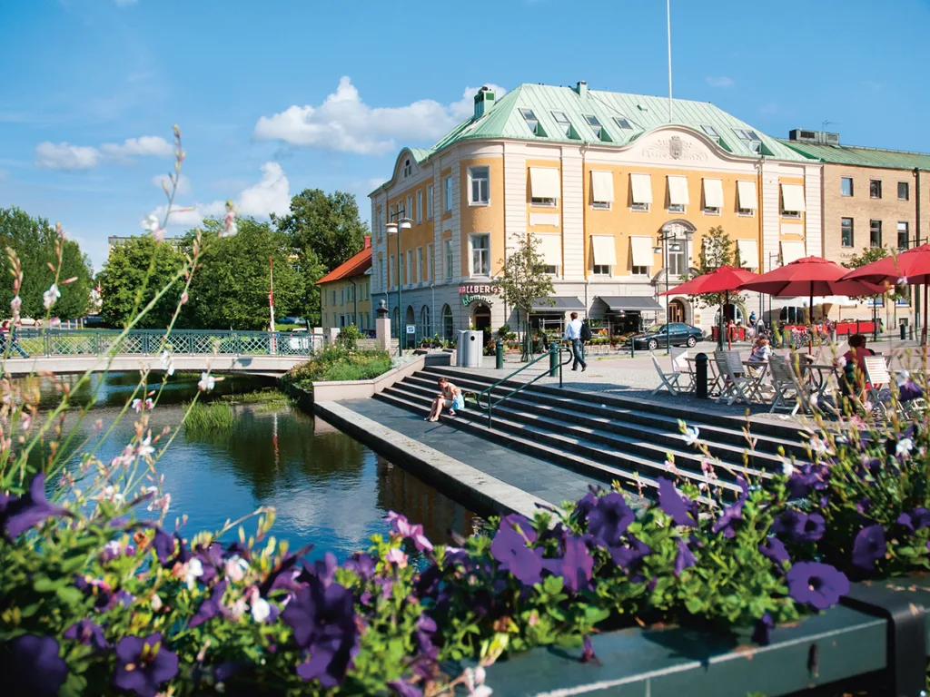 Ett naturskönt torg vid floden med lila blommor i förgrunden, uteserveringar med röda parasoller och en historisk gul byggnad under en ljusblå himmel. Människor kopplar av vid vattnet och promenerar nära bron.