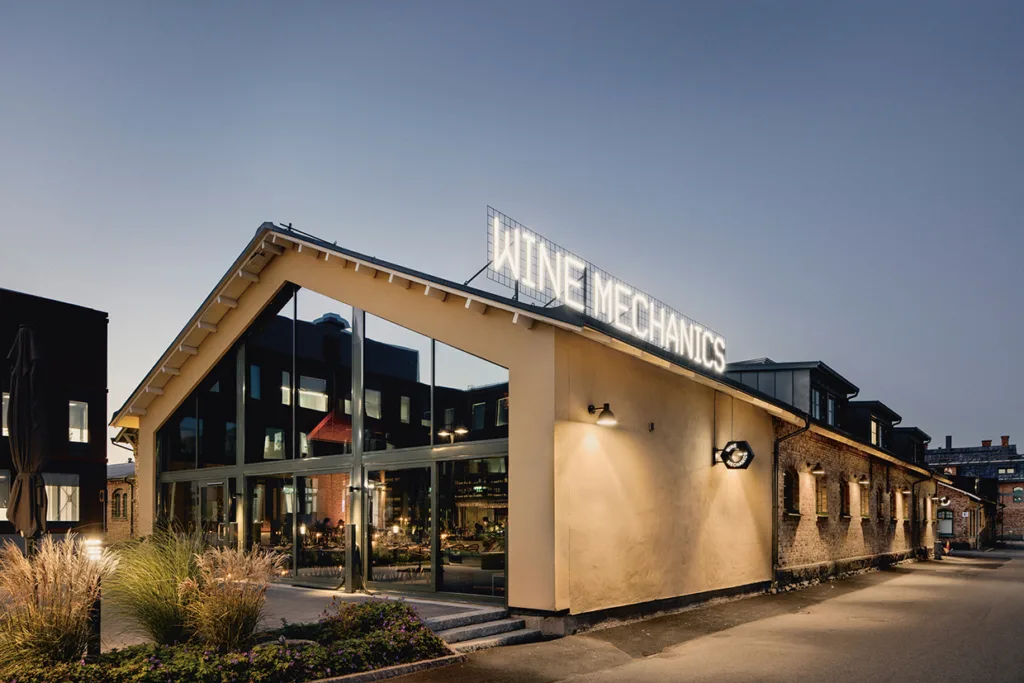 En modern byggnad med stora glasfönster och en skylt på taket med texten WINE MECHANICS, fotograferad i skymningen med synliga varma innerlampor och planteringar framför.