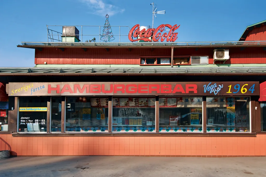 En hamburgerbar i retrostil med stor HAMBURGERBAR-skylt, Coca-Cola-skylt på taket och 1964 målat på byggnaden. I fönstren finns menyer och exteriören är röd med gröna och turkosa inslag.