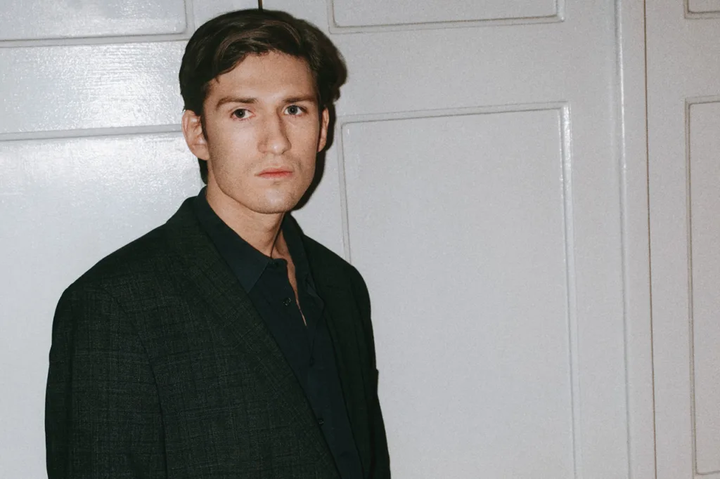 Isac Calmroth med kort brunt hår, klädd i mörk kavaj och skjorta, står framför en vitmålad vägg och tittar lite åt sidan med ett neutralt ansiktsuttryck.