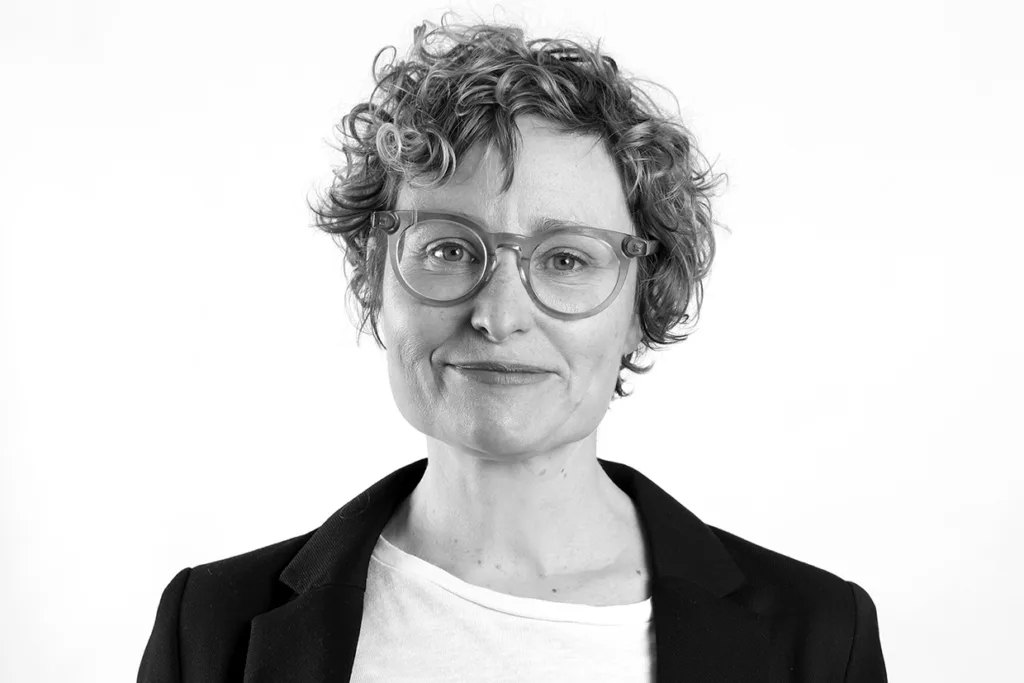 Ylva Ågren med kort, lockigt hår och glasögon, klädd i en mörk kavaj över en ljus skjorta, ler lätt medan han tittar in i kameran mot en vit bakgrund.
