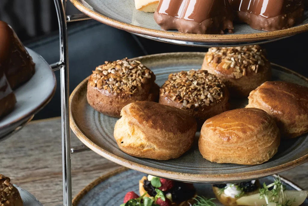 Närbild på en serveringsbricka i flera nivåer med gyllene scones och bakverk med nötter på en nivå och chokladglaserade desserter på den övre nivån, med mer mat synlig på en tallrik nedanför.