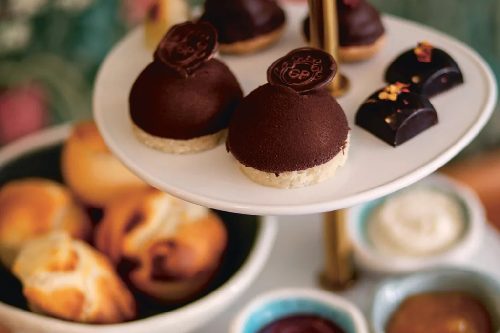 Närbild på ett dessertställ i flera nivåer med kupoler av chokladmousse med chokladmedaljonger, glansiga chokladpraliner och en lägre tallrik med olika bakverk och små skålar med sylt och grädde.